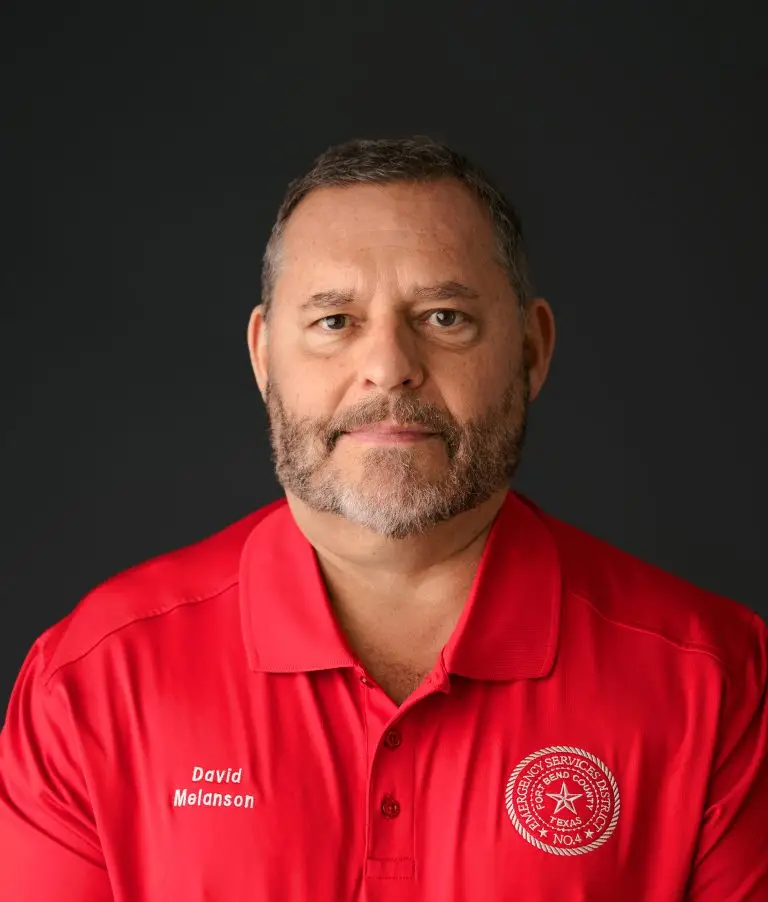 A man with a beard wearing a red polo shirt with a logo on the right side and the name "David Melanson" embroidered on the left side. He is posing against a dark background and looking directly at the camera with a neutral expression.