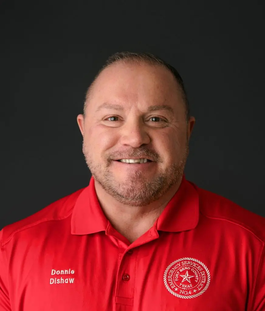 A man with short hair and a beard smiles at the camera. He is wearing a red collared shirt with an embroidered Texas state emblem on the right and "Donnie Dishaw" on the left. The background is plain and dark.