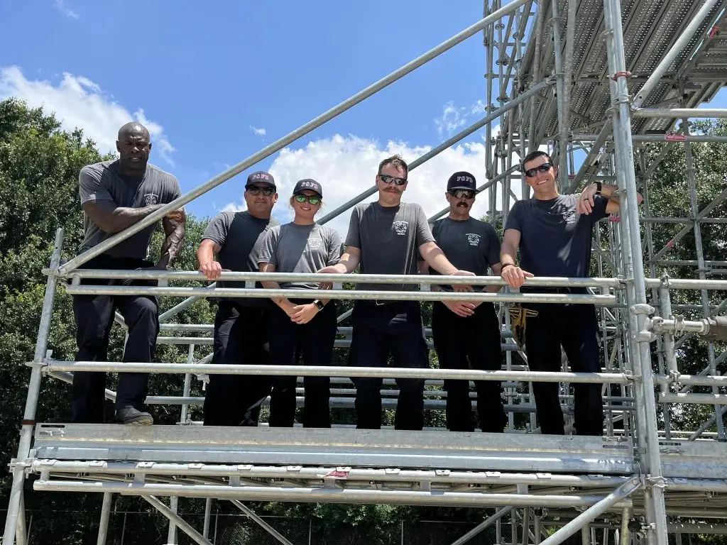 Six construction workers stand on metal scaffolding outdoors, posing for a group photo. They wear matching black shirts, some with hats and sunglasses. The background includes trees and a blue sky.