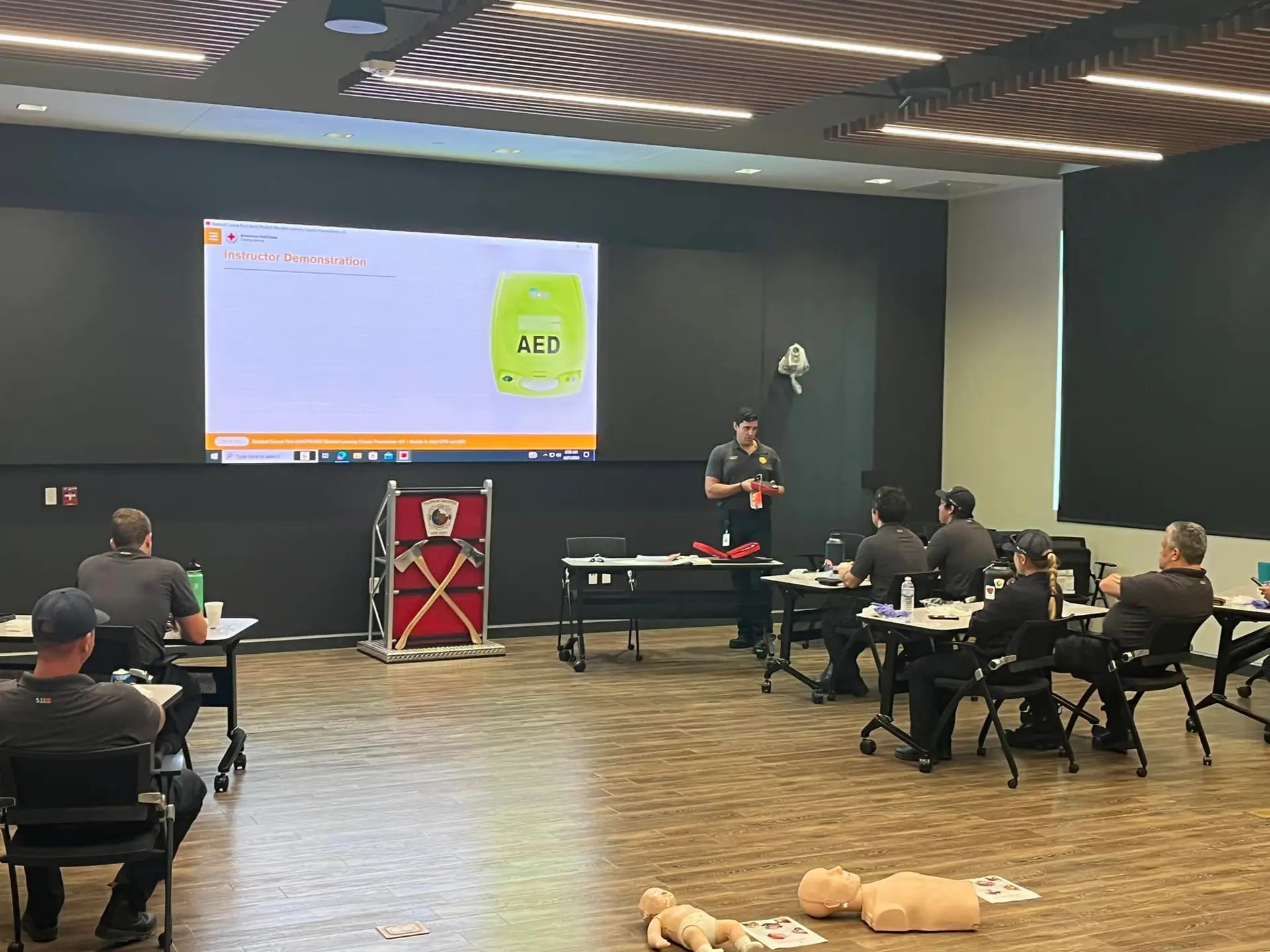 A man stands at the front of a classroom giving a presentation on an AED (Automated External Defibrillator) device. Six other people are seated looking at the presentation screen. Mannequins for CPR training are on the floor near the presenter.