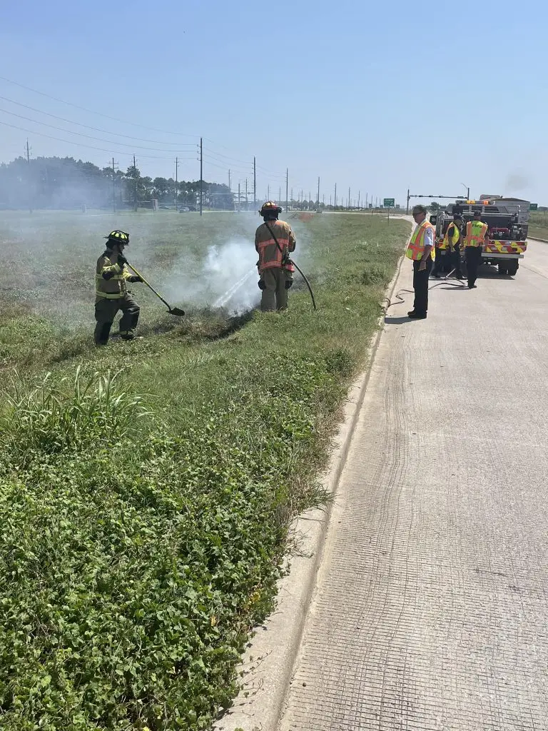 Two firefighters in gear battle a small grass fire beside a road. One sprays water while the other uses a tool to control the flames. Two emergency personnel in high-visibility vests stand near fire trucks parked on the roadside.