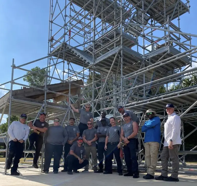 A group of 14 people posing in front of a large scaffolding structure. Some are standing, and some are kneeling. They are dressed in various casual work attire and safety gear, suggesting a team engaged in construction or maintenance work. The sky is clear and sunny.