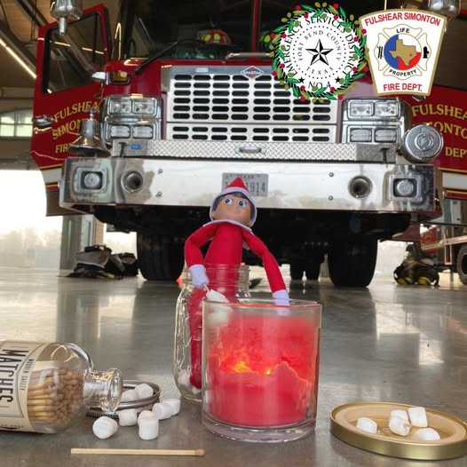 A festive toy elf is positioned in front of a red fire truck inside a fire station. In the foreground, there's a lit red candle, a jar of matches, matches scattered around, and mini marshmallows. Fire department logos are visible above.